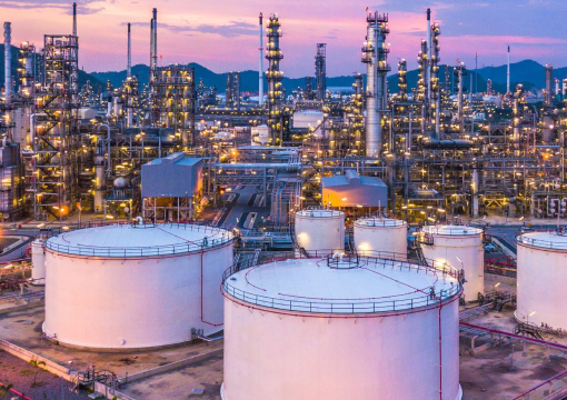 An industrial oil refinery at dusk with large white storage tanks in the foreground and numerous illuminated metal towers and pipes in the background, set against a purple and pink sky.